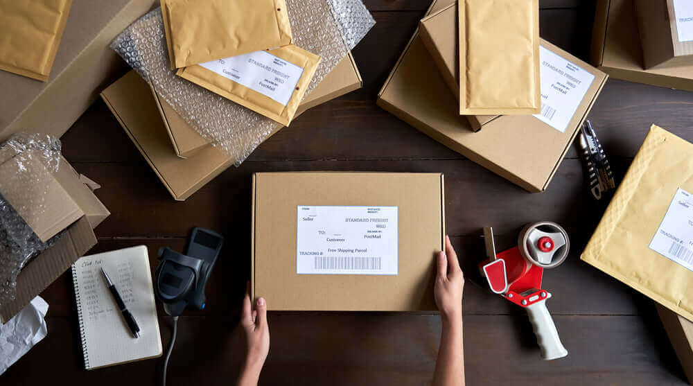 Hands holding a shipping box surrounded by packed parcels and shipping materials on a wooden table.