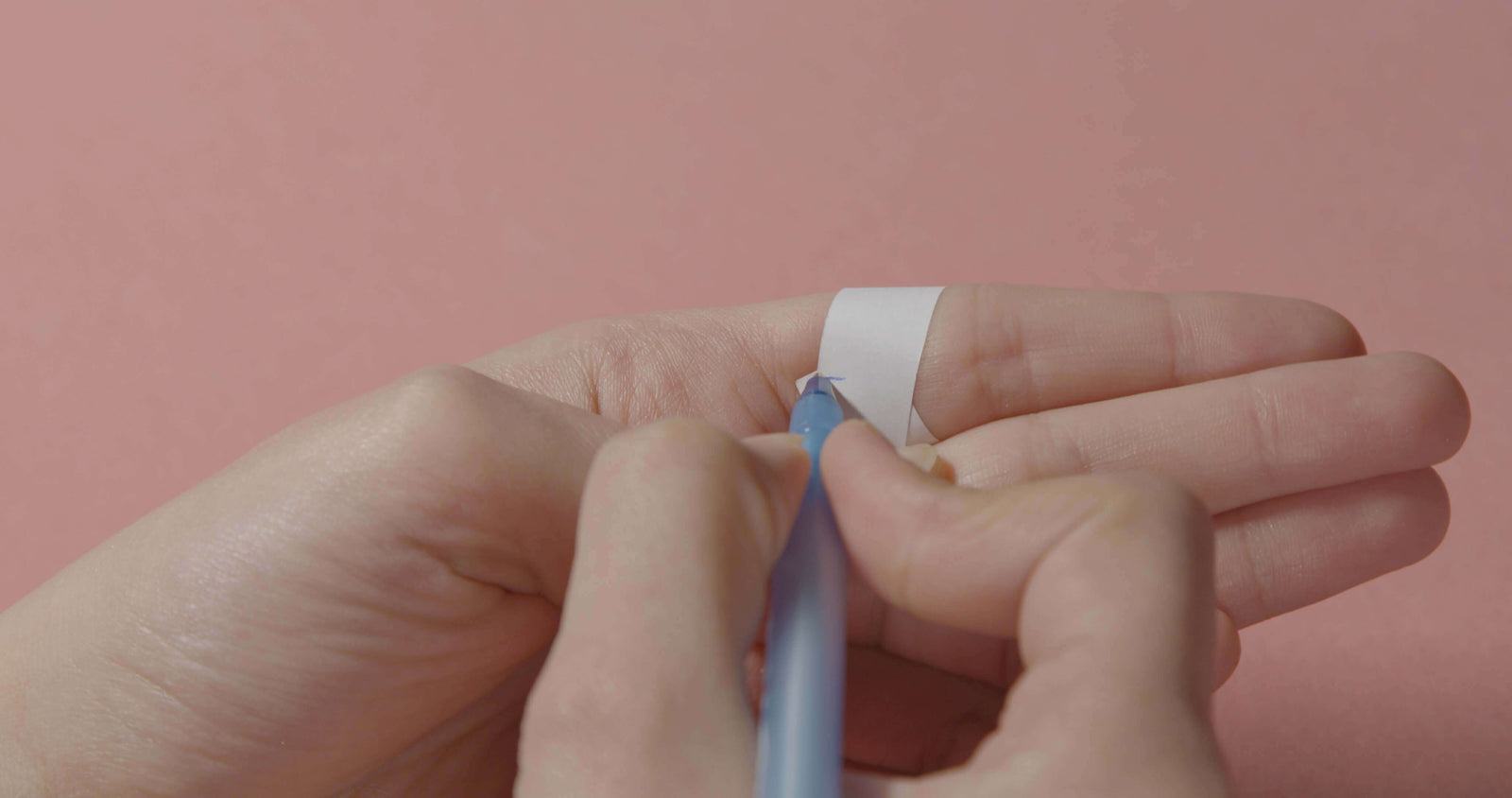 Person measuring ring size at home using a paper strip and pen on a light pink background.