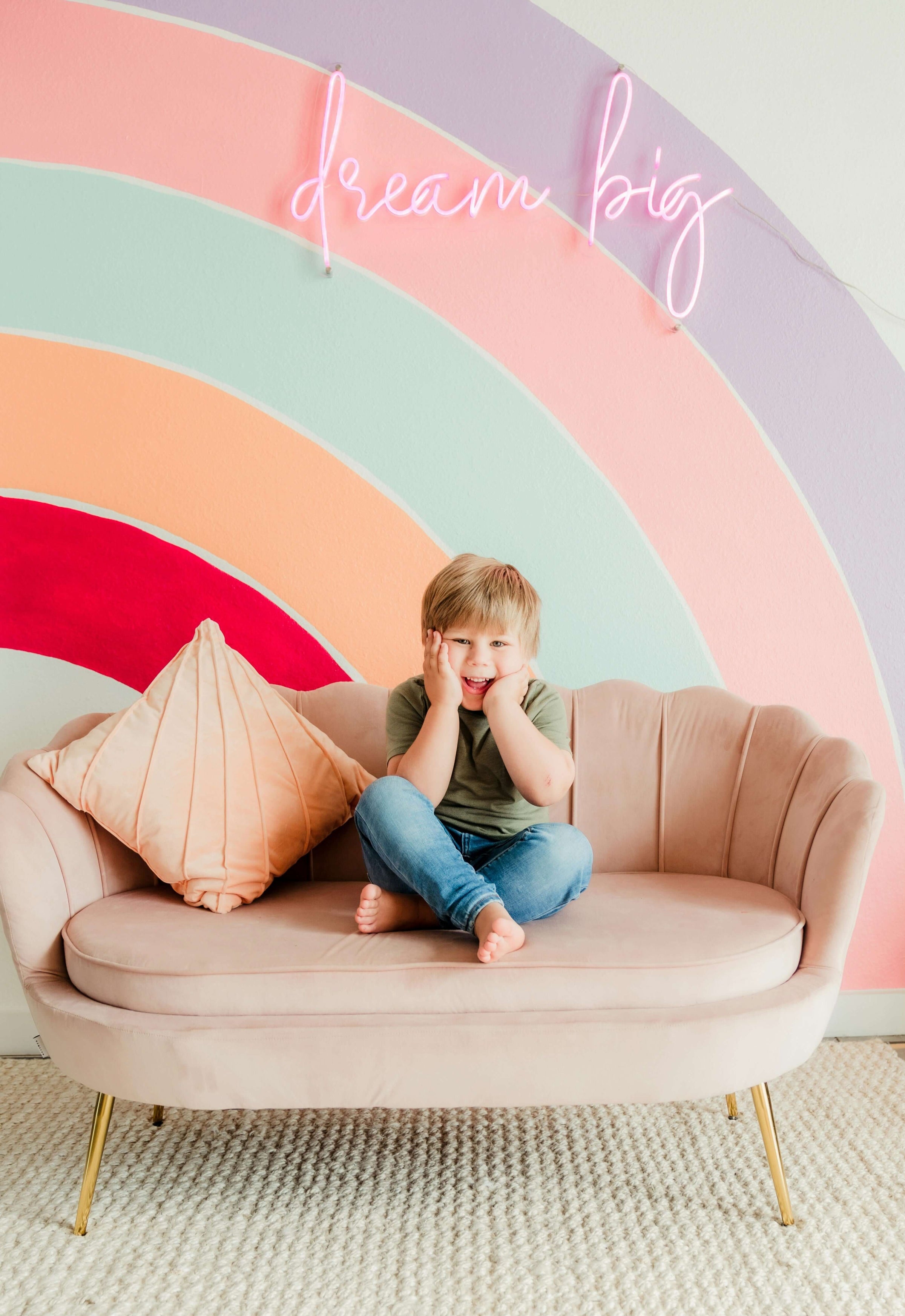 Child sitting on a pink couch in front of a colorful rainbow wall with "dream big" text, showcasing fun, affordable kids' room decor.