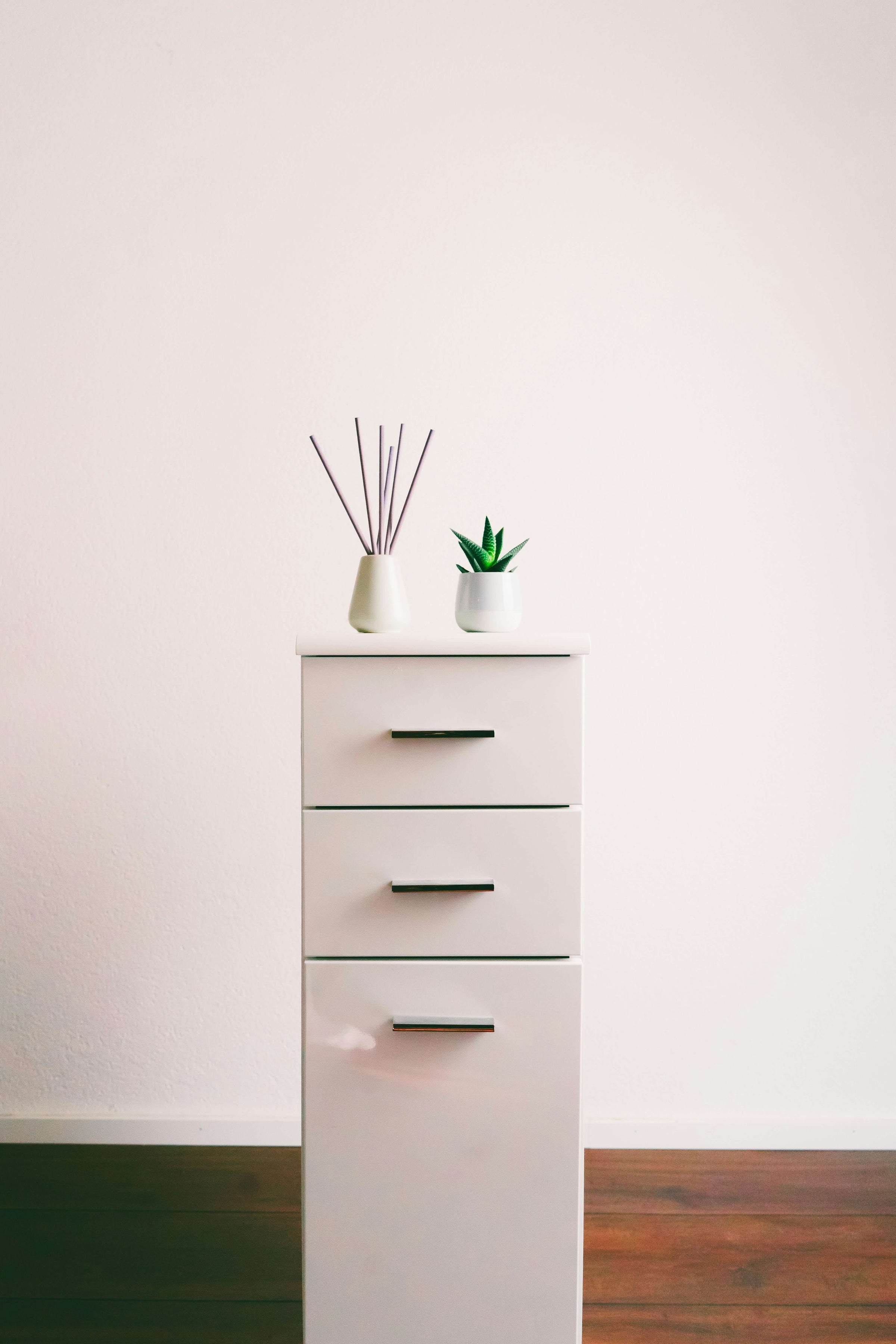 White office storage cabinet with three drawers, topped with a small plant and diffuser, on a wooden floor.