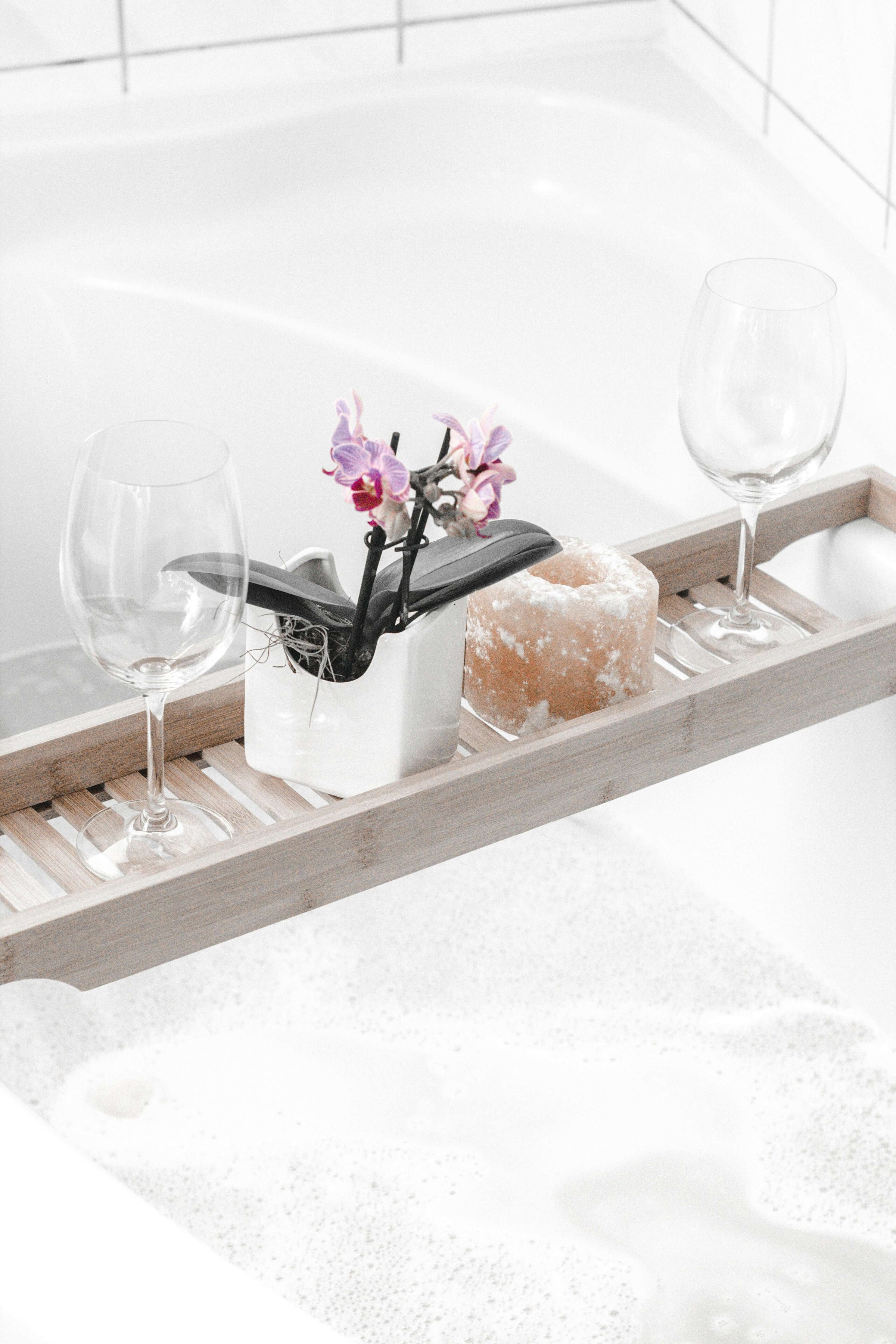 Elegant wooden bath tray with wine glasses, a white vase with flowers, and a candle, enhancing bathroom décor with affordable style.