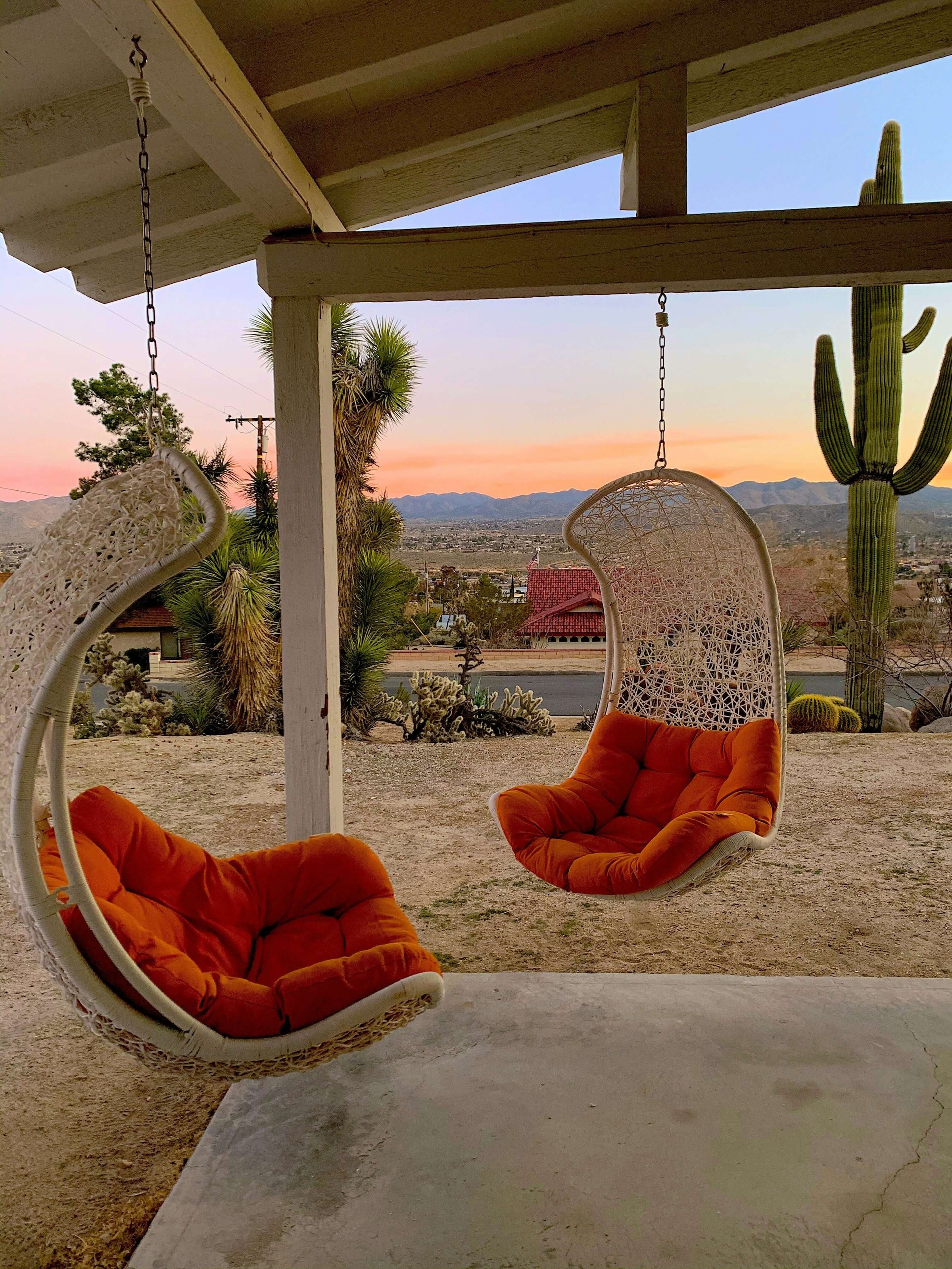 Cozy outdoor hanging chairs with orange cushions under patio roof at sunset in a desert landscape.