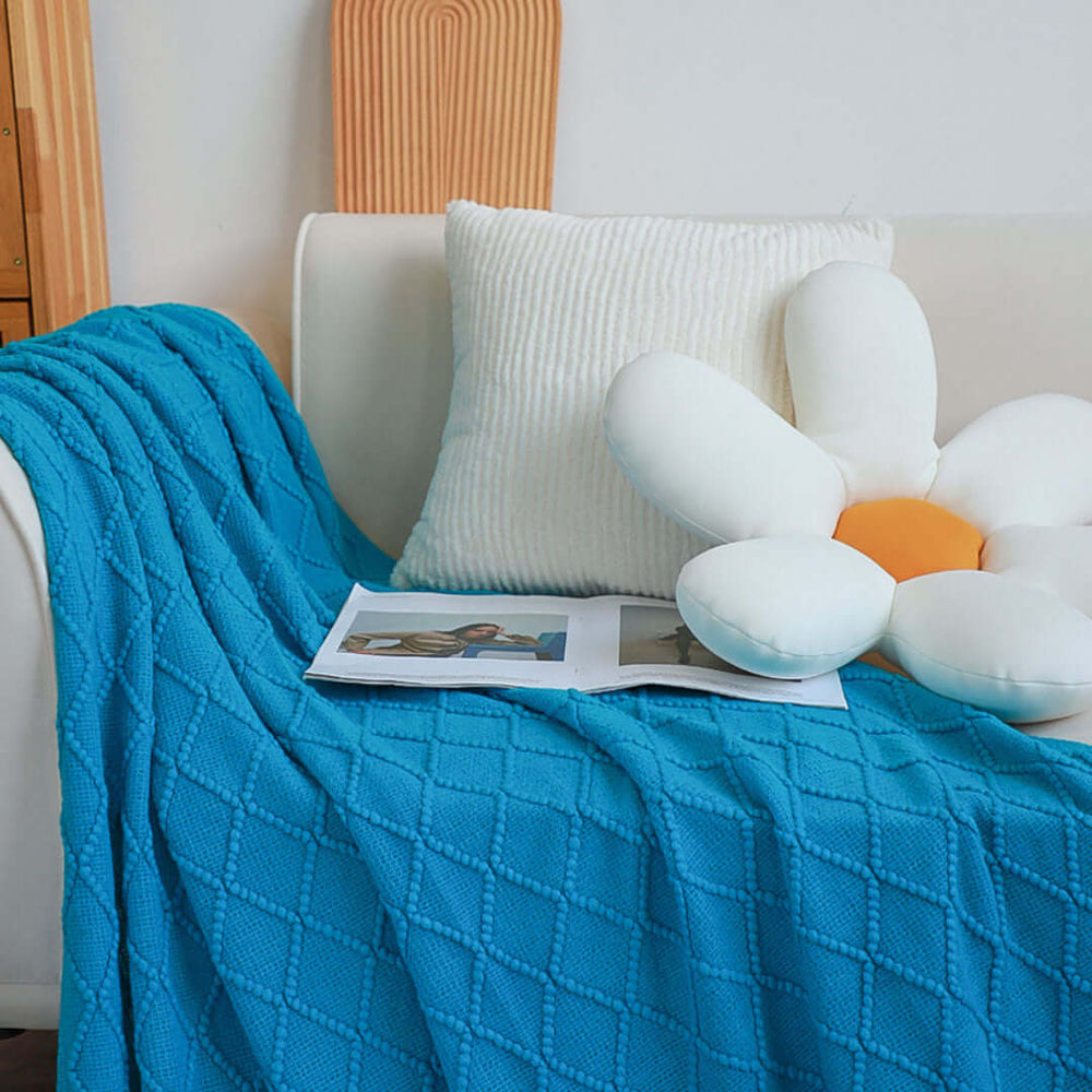 Cozy white couch with a blue textured blanket, white flower-shaped cushion, and a magazine, showcasing affordable quality homewares and value furniture.