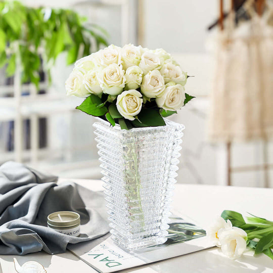 Crystal vase with a bouquet of white roses on a table, surrounded by homewares and decor in a well-lit room.