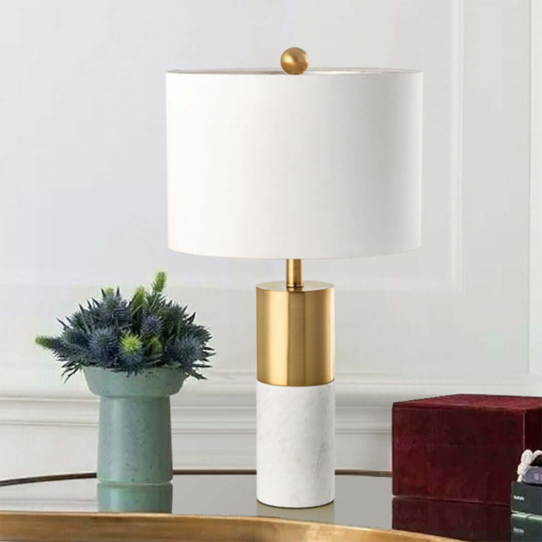 Modern affordable homeware table lamp with gold accents sitting on a glass table, next to a potted plant and decorative box.