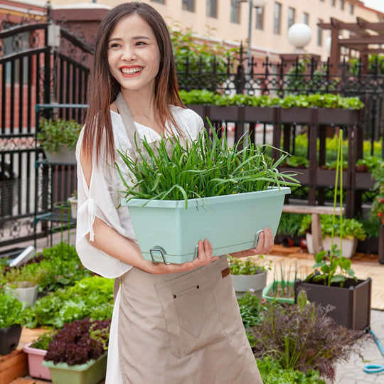 Woman smiling while holding a planter filled with fresh green plants in an outdoor garden area.