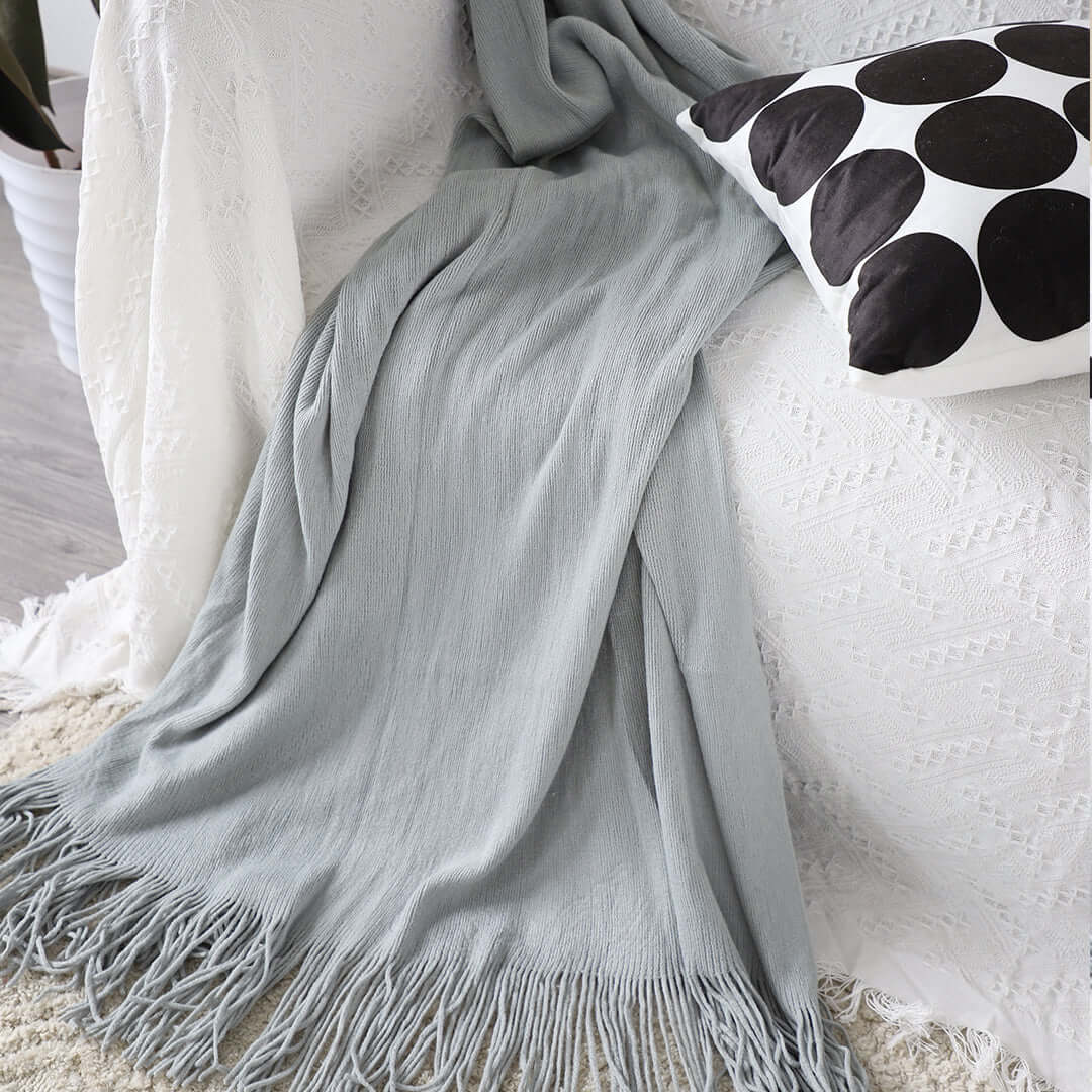 Affordable quality homewares featuring a stylish gray throw blanket with fringe and a decorative black and white pillow on a white sofa.