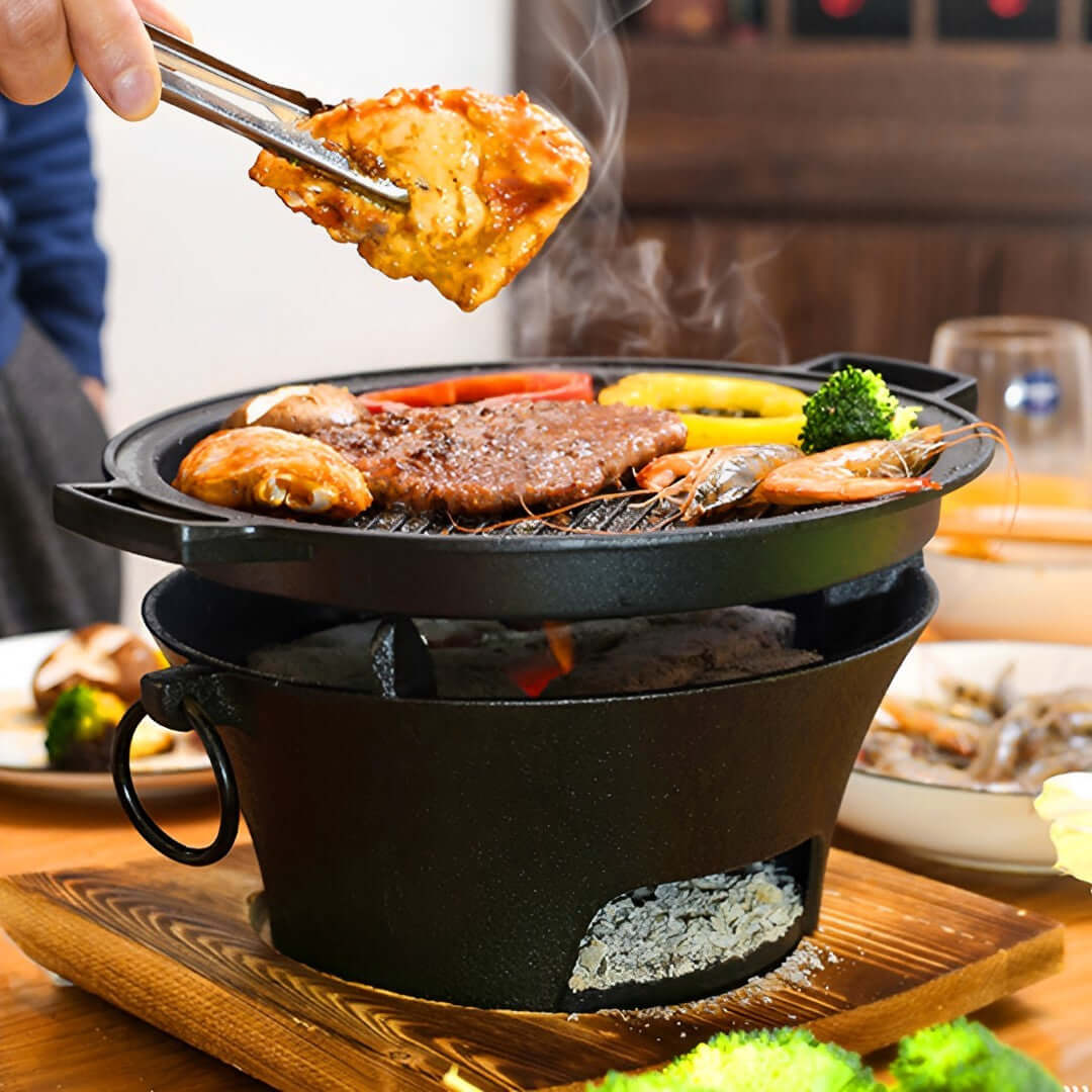 Person grilling meat and vegetables on a small charcoal grill at a dining table.