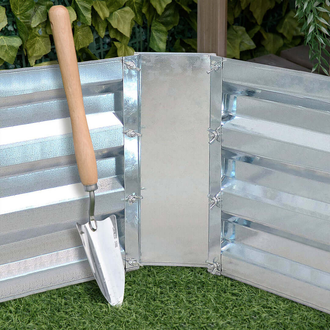 Metal garden trowel resting against a corrugated metal garden bed on green grass with lush foliage in the background, showcasing durable gardening tools