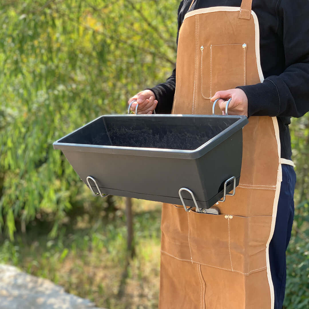 Person holding a black rectangular planter with metal hooks while wearing a brown apron outdoors.