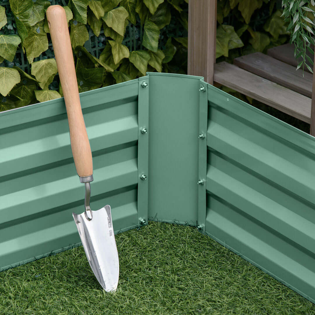 Garden trowel leaning against a green metal-raised garden bed with artificial grass and green plants in the background.