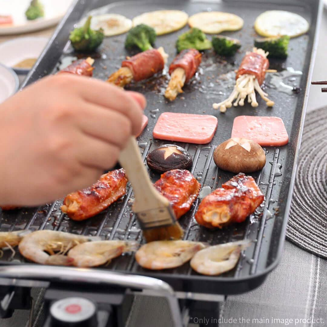 Hand brushing sauce on grilled food including shrimp, mushrooms, broccoli, and bacon-wrapped vegetables on a grill.