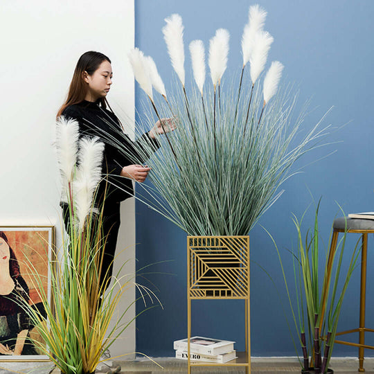 Woman arranging tall artificial grass plant in a stylish metal planter next to value homeware pieces including framed art and indoor plants.