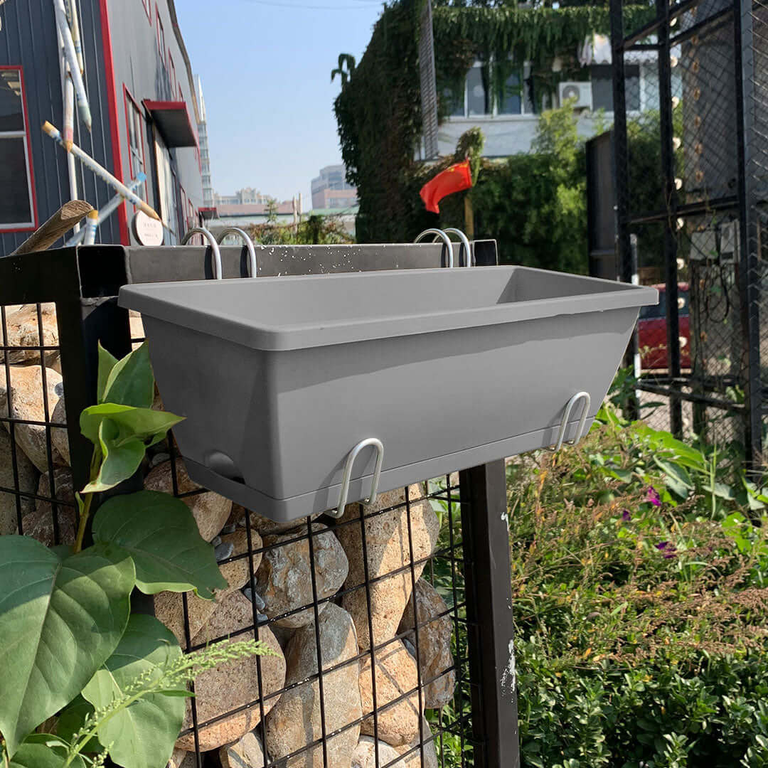 Affordable grey planter box attached to a metal fence in a garden setting with lush green plants and urban background.