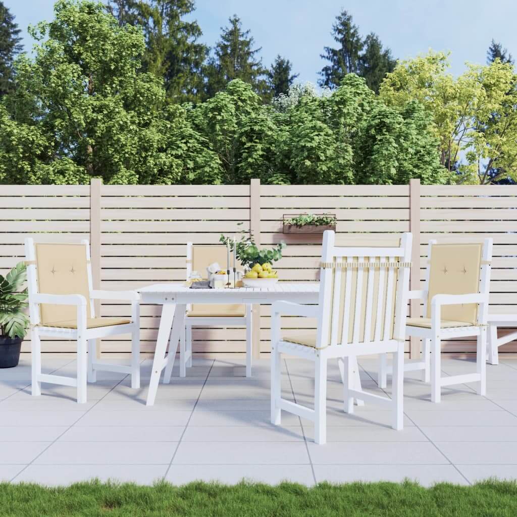 Outdoor dining set with white table and chairs on patio, beige cushions, fenced garden with lush green trees in the background.