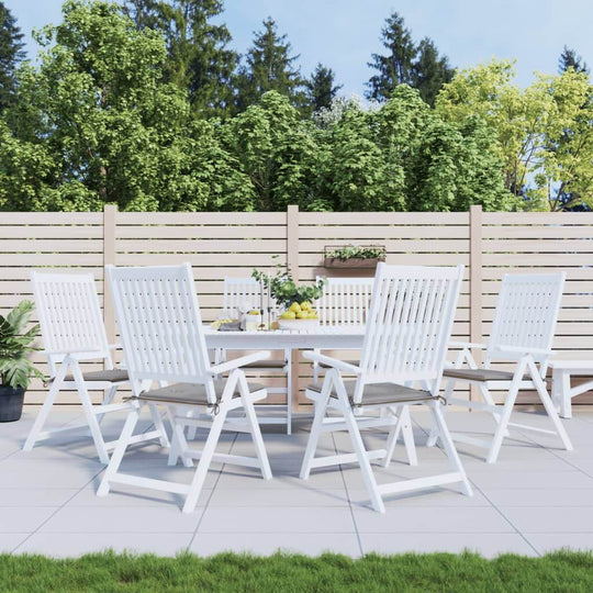 Outdoor dining set with white wooden chairs and table on a patio, surrounded by greenery and a wooden fence.