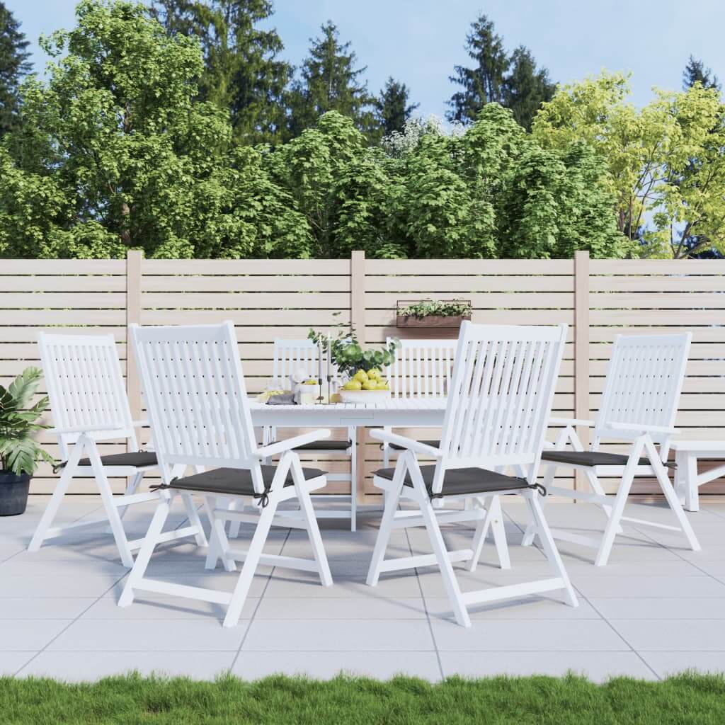 Outdoor dining area with white wooden chairs and a round table set against a wooden fence and lush green trees in the background.