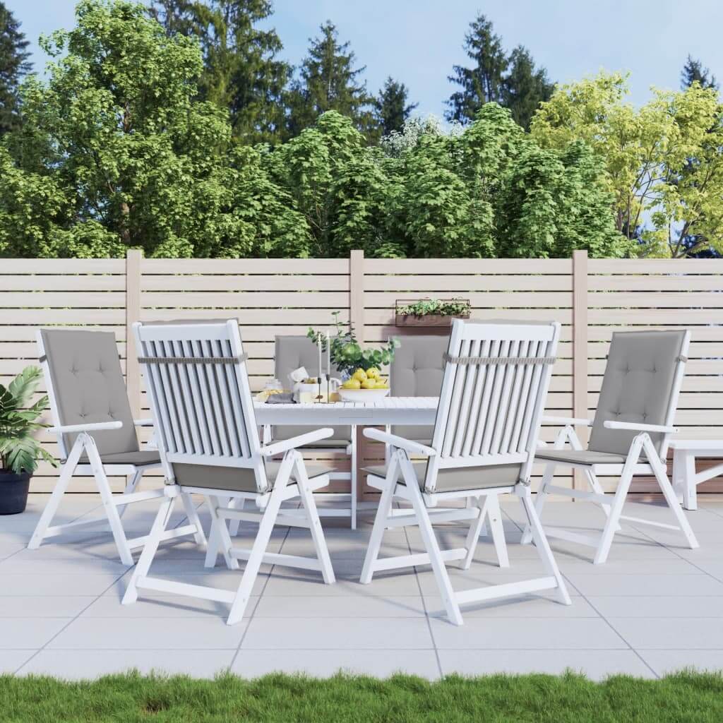 Outdoor patio with white chairs featuring grey highback cushions and a round dining table, set in a garden with greenery and a wooden fence in the background.