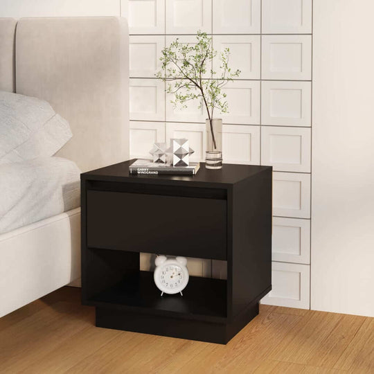 Black bedside cabinet with drawer and open compartment, next to a bed, featuring a clock and decorative items on top.