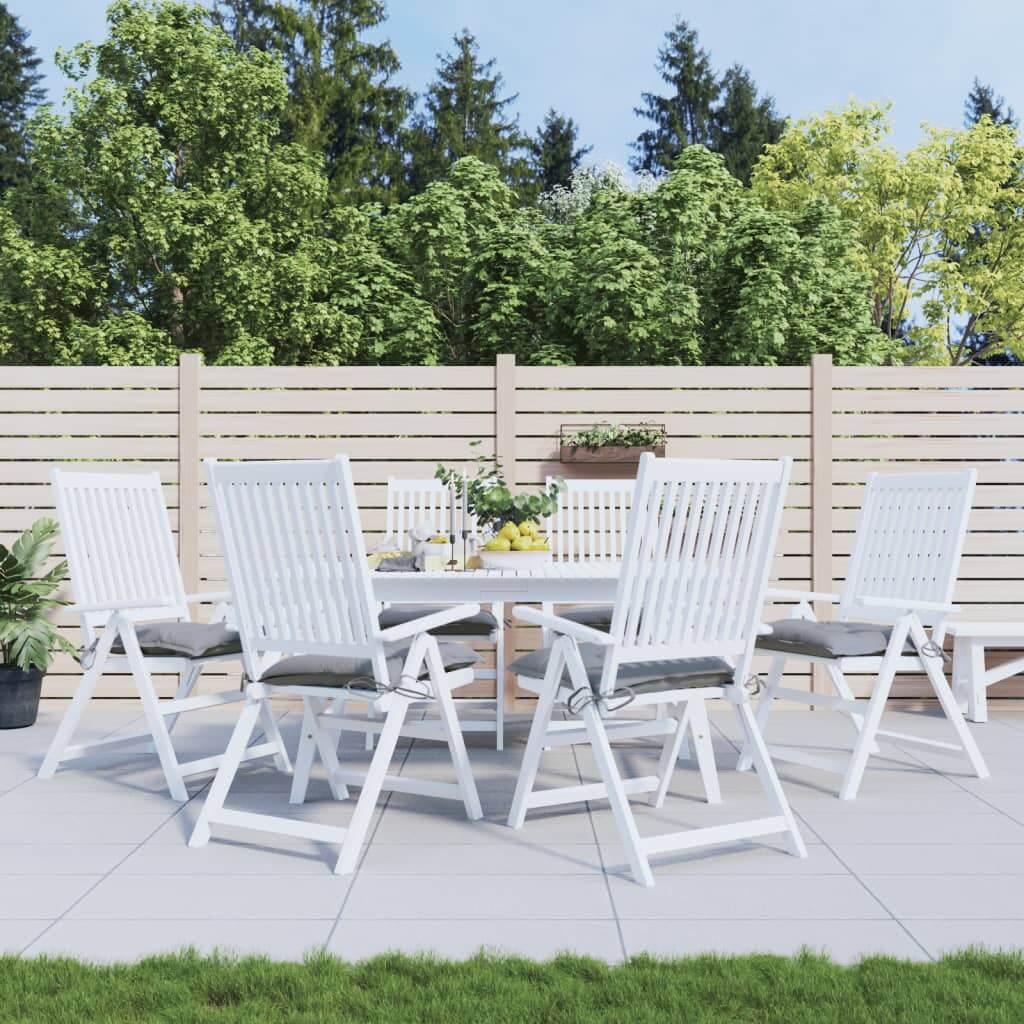 White outdoor dining set with grey chair cushions in a patio space, surrounded by greenery and wood fencing.