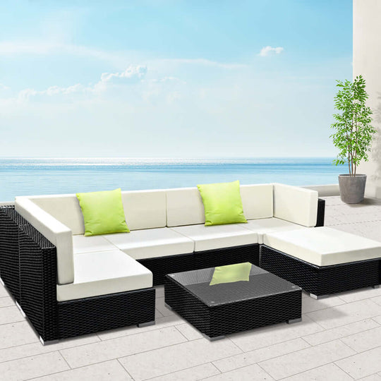 Gardeon 7-Piece Outdoor Sofa Set with black wicker, white cushions, and green accents by the sea for a luxurious lounge experience.