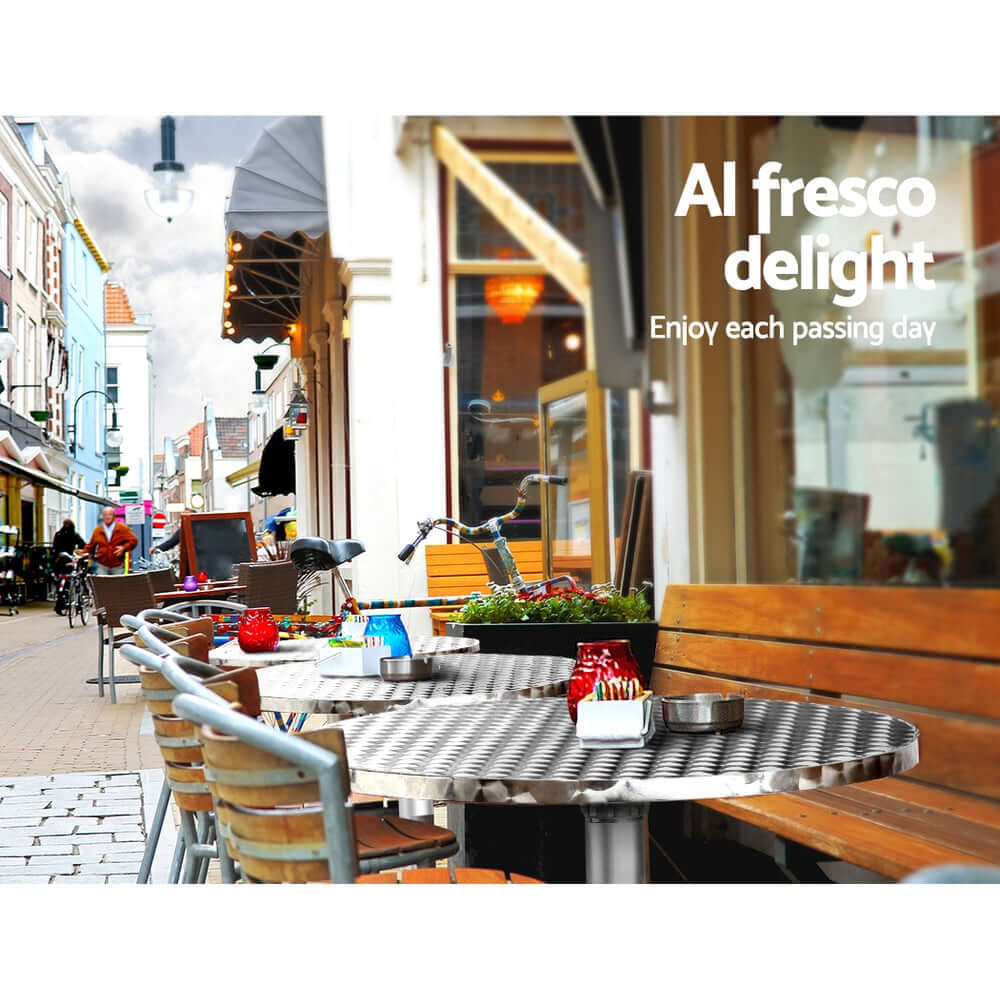 Outdoor bar table set for al fresco dining, featuring stainless steel top and stylish seating in a vibrant street setting.