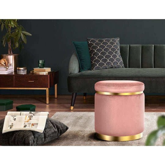 Elegant round pink velvet ottoman footstool with gold trim in a stylish lounge setting.