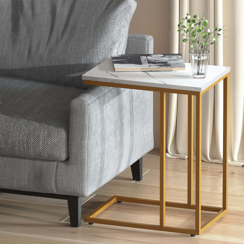 Modern Artiss Coffee Table with marble finish and gold metal frame next to a gray sofa with a book and plant on top.