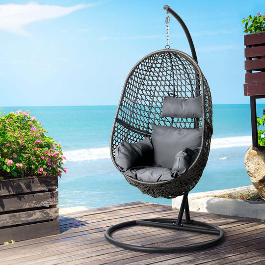 Gardeon outdoor egg swing chair in black wicker rattan with cushion on a patio by the sea, perfect for relaxation.