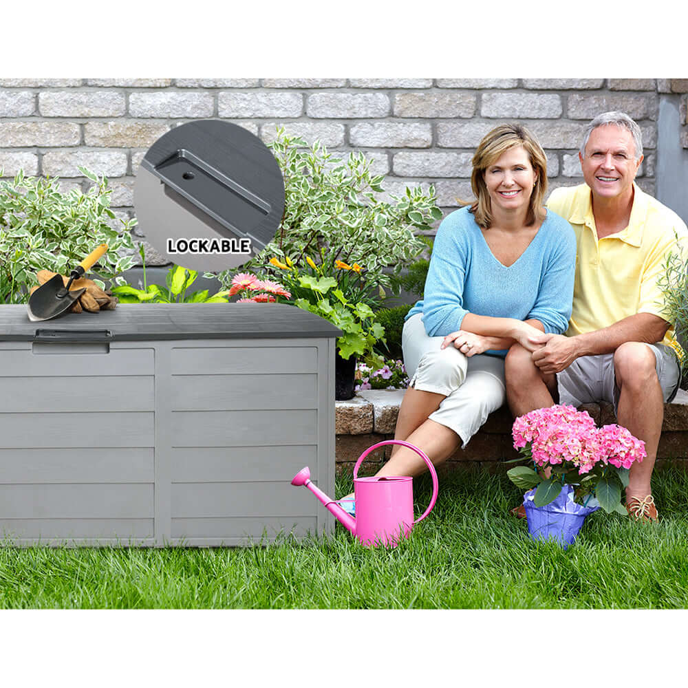 A happy couple sitting by a grey Gardeon Outdoor Storage Box, showcasing its lockable feature and garden setting.