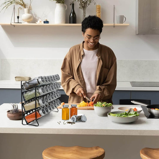 Woman preparing food with a 4-tier spice rack organizer featuring 28 spice jars on a modern kitchen countertop.