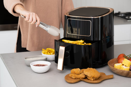 Person using 7L Digital Air Fryer (Black) to cook fries, alongside a meal of fried chicken and vegetables on a kitchen counter.