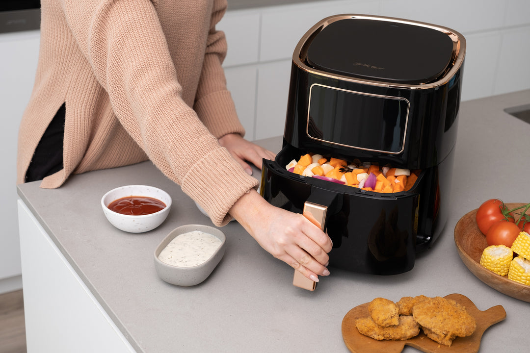 Person using 7L Digital Air Fryer in black to cook vegetables, with bowls of sauces and fresh ingredients on a kitchen countertop.