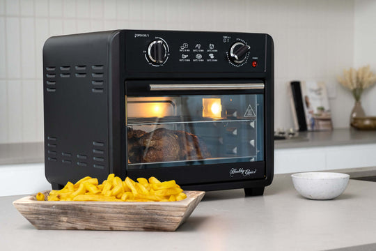 23L black air fryer oven with baking tray, wire rack, and pan handle, cooking a meal in a modern kitchen, next to a dish of crispy fries.