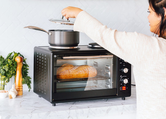 Woman using portable oven with rotisserie cooking and hot plate, baking bread alongside vegetables and spices on kitchen counter.