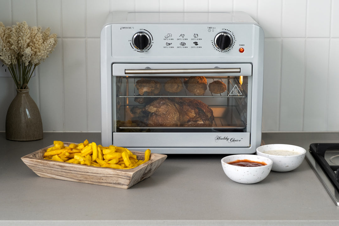 23L Air Fryer Oven in silver on a kitchen countertop cooking chicken, accompanied by a wooden tray of fries and two bowls of dips.