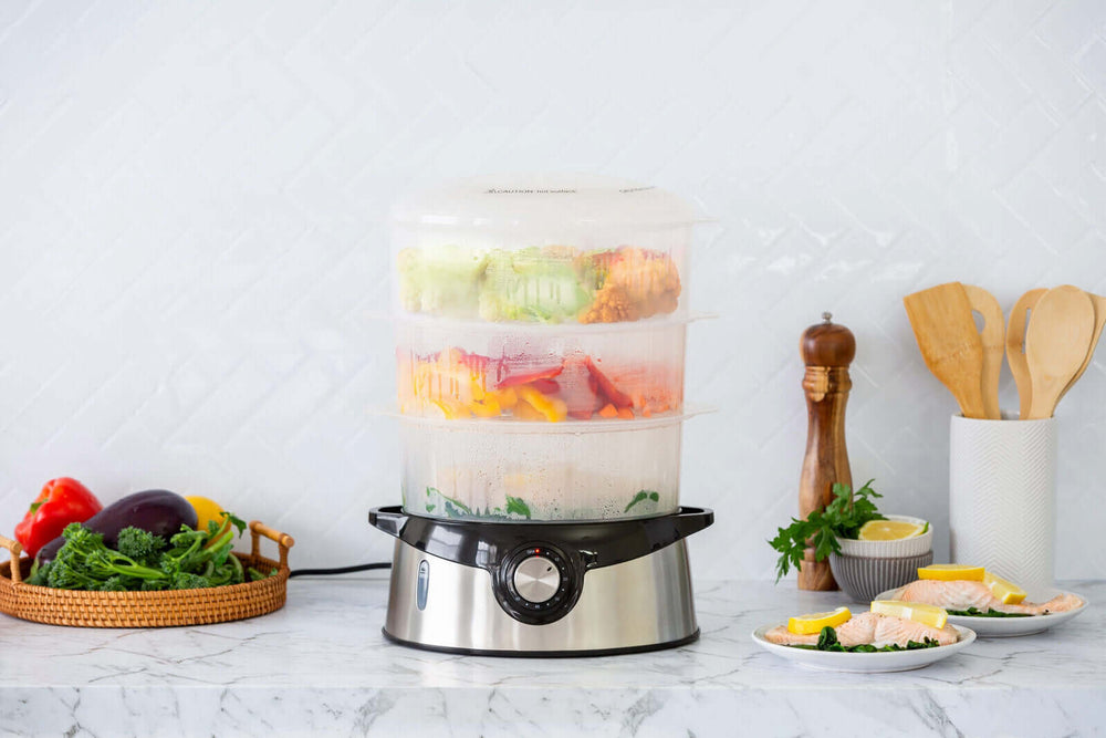 3 tier food steamer with stainless steel base steaming colorful vegetables and fish on a kitchen countertop.