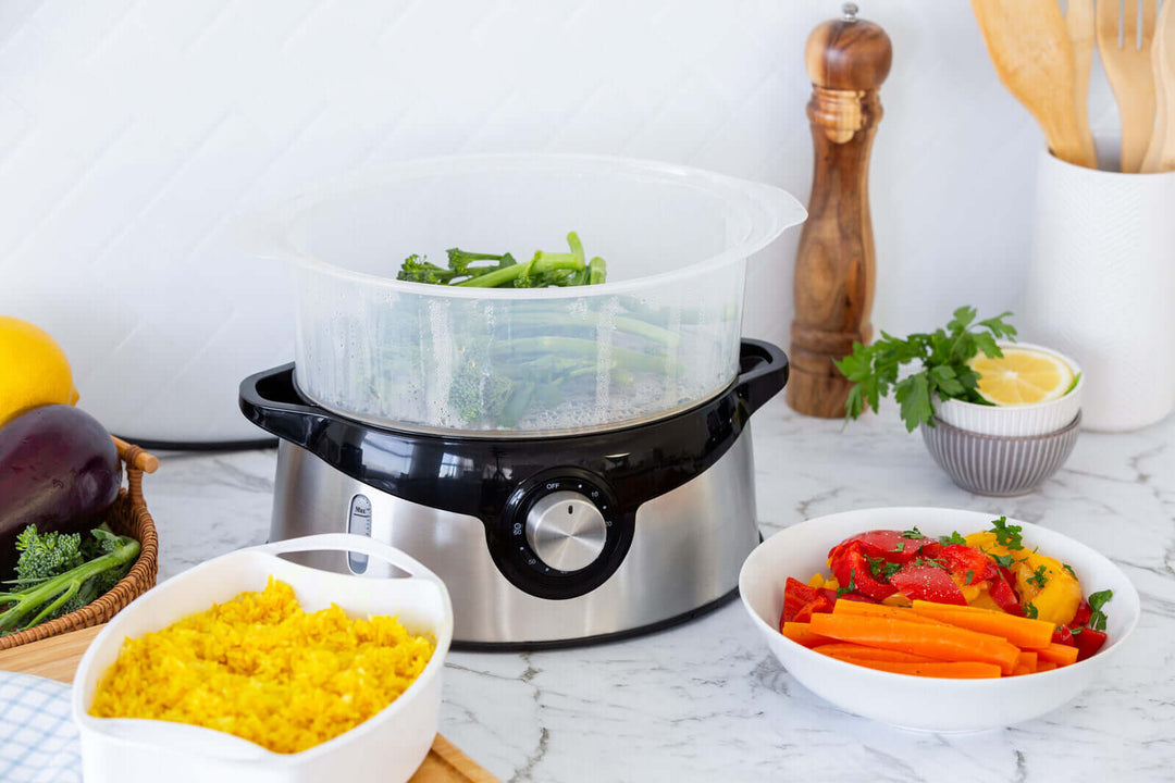 3 tier food steamer with stainless steel base, steaming fresh vegetables, surrounded by healthy dishes on a marble countertop.