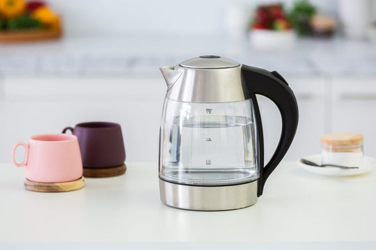 Glass kettle with black handle and 360 degrees rotational base on a kitchen countertop, next to two colorful mugs and kitchen accessories.