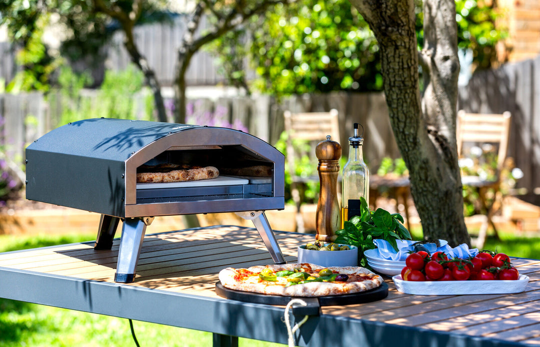 Outdoor scene with Compact and Portable 12" Electric Pizza Oven on table, cooking pizza surrounded by fresh ingredients and kitchenware.