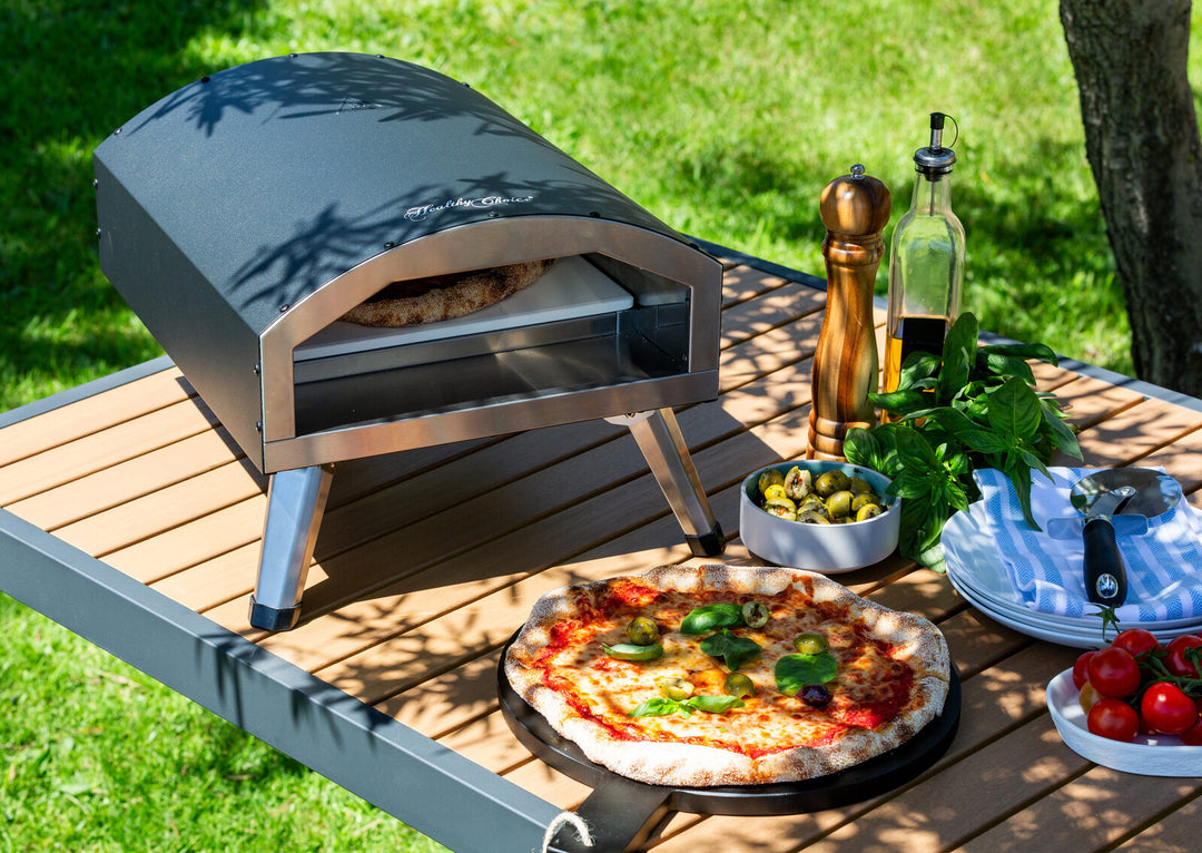 Compact and portable 12" outdoor electric pizza oven on a table with a freshly baked pizza, vegetables, and condiments in a garden setting.