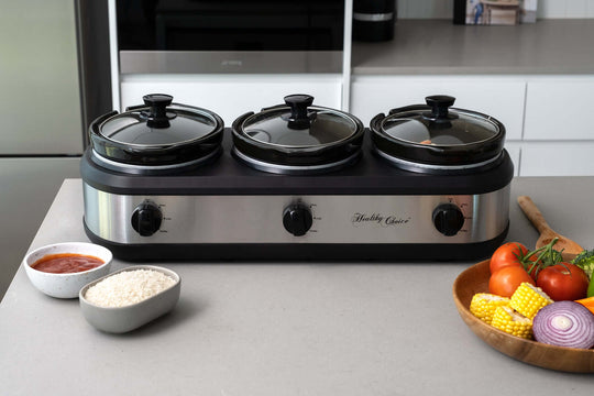 3-Pot Slow Cooker with three glass lids, adjustable knobs, and bowls of ingredients on a kitchen countertop.