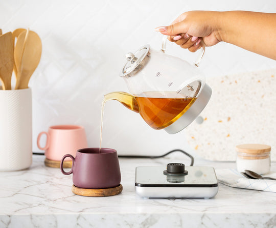 Pouring tea from a 1.2L digital glass kettle with infuser into a purple mug on a marble countertop with wooden utensils and a digital base.