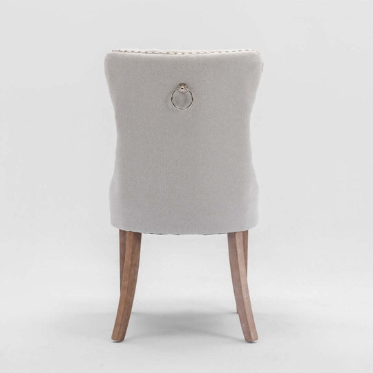 Back view of a beige button-tufted dining chair with stud trim and wooden legs, showcasing elegant design details.