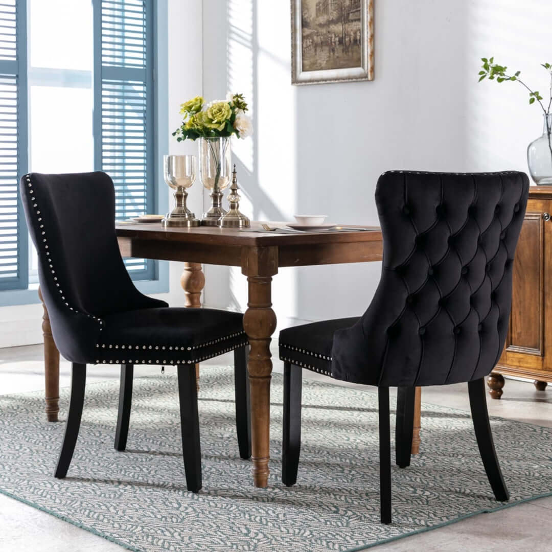 Black velvet upholstered dining chairs with tufted design and studs trim around a wooden dining table set for two, in a modern elegant kitchen.