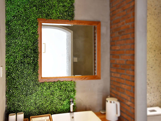 Lush green artificial plant wall panel in a modern bathroom setting with a wooden mirror and brick decor.