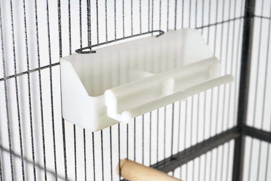 Close-up of a white food or water dish inside a large bird cage with wooden perch.