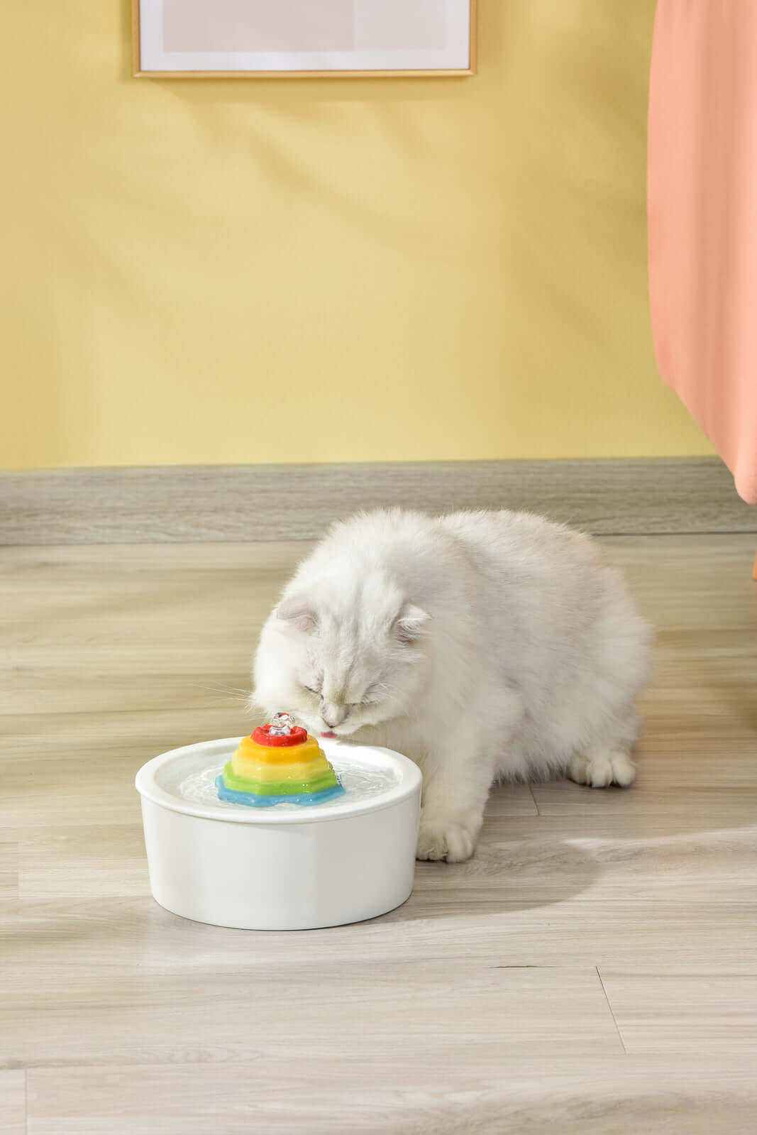 White cat drinking from a colorful ceramic electric pet water fountain on a wooden floor.