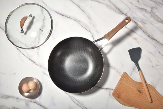 Non-stick stainless steel Noka wok with lid, spatula, and eggs on marble countertop, showcasing quality and affordability.