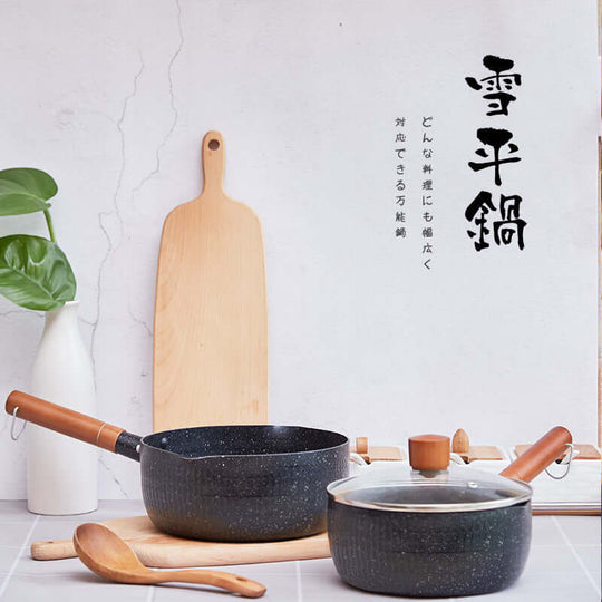 Justcook black aluminum Xueping pot with wooden handle and lid, alongside a wooden spatula and plant on a kitchen countertop.